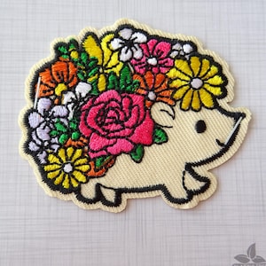 May include: Embroidered iron-on patch of a white hedgehog with a crown of colourful flowers, including pink roses, yellow daisies, and orange and purple flowers.