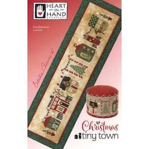 May include: A cross-stitch pattern featuring a Christmas tiny town design. The pattern includes a long, rectangular piece with stitched houses and trees, a red pincushion, and the text "Christmas tiny town."