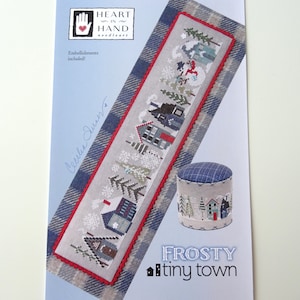 May include: A cross-stitch pattern depicting a winter scene with miniature houses, snow-covered trees, and a snowman. The design is framed with a red border and includes the text "Frosty tiny town". A small, matching round pincushion is also shown.