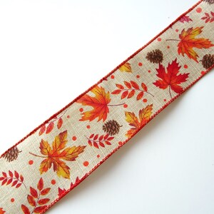 May include: A beige ribbon with a red edge features a repeating pattern of orange, yellow, and red leaves, along with small brown pinecones and red dots.