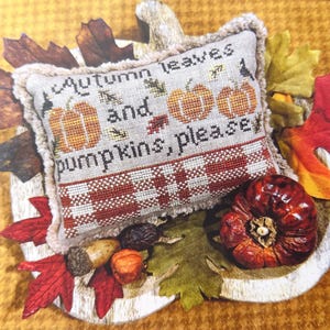May include: A decorative pillow embroidered with the words "Autumn leaves and pumpkins, please". The pillow features images of pumpkins and is surrounded by autumn leaves and a small pumpkin. The pillow has a red and white checkered pattern.