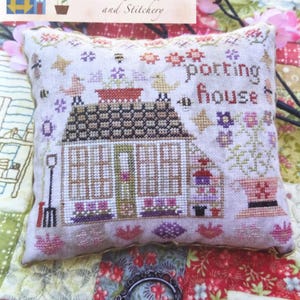May include: A cross-stitch cushion featuring a small house with a red roof and the words "potting house" embroidered on the front. The cushion is decorated with flowers, birds, and bees.