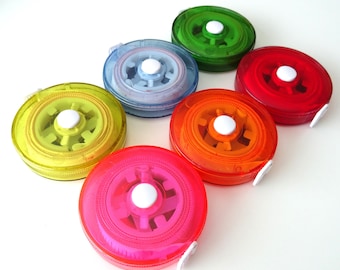 May include: A collection of colorful, transparent tape measures in shades of yellow, blue, green, orange, red, and pink. Each tape measure has a white center and a white pull tab. The tape measures are arranged on a white surface.