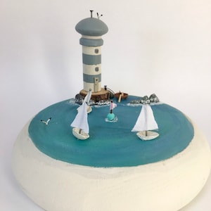 Puede incluir: Un faro en miniatura con una torre rayada de gris y blanco se alza sobre una base redonda con una escena de océano pintada de azul. Dos pequeños veleros blancos están en el agua, y una pequeña boya rosa y blanca flota en el agua.