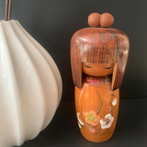 Könnte beinhalten: Japanische Kokeshi-Puppe aus Holz mit braunem Kopf und orangefarbenem Körper. Die Puppe hat ein einfaches Gesicht und ist mit floralen Designs in Weiß, Rosa und Grün verziert. Daneben eine weiße Vase mit geschwungenem Design.