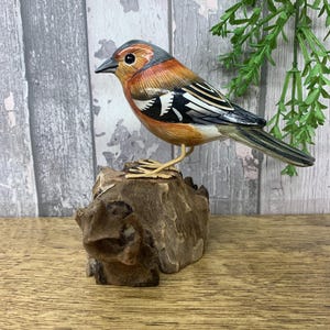 Puede incluir: Escultura de pájaro de madera tallada a mano. El ave presenta un diseño detallado con plumas naranjas, marrones, negras y blancas. Está posada sobre un trozo de madera natural, mostrando la intrincada artesanía.