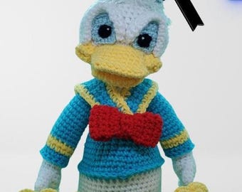 Op de afbeelding: Een handgemaakte gehaakte pop van Donald Duck. De pop heeft een blauwe matrozenhoed, een wit gezicht met een gele snavel en een blauw-gele trui met een rode vlinderdas. De voeten van de pop zijn geel en hij zit.
