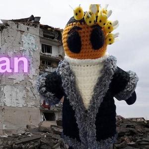 May include: A crocheted bee plush toy wearing a blue coat with a gray fur trim. The toy is standing in front of a destroyed building.