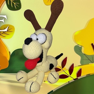 May include: A crochet pattern for making a plush Odie dog from the comic strip "Beetle Bailey". The pattern is available as a PDF download. The image shows a crocheted Odie dog with a red tongue sticking out, standing in front of a cartoon background of fall leaves.