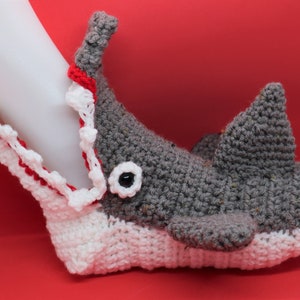 May include: A gray and white crocheted shark slipper with a red mouth and a white eye. The slipper is open and appears to be biting down on a white object.