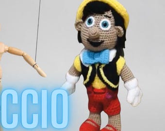 Puede incluir: Un títere de crochet de Pinocho, un personaje clásico de Disney. El títere lleva un sombrero amarillo, un chaleco negro con ribete amarillo, pantalones rojos y zapatos rojos. El títere tiene una pajarita azul y una sonrisa amigable.