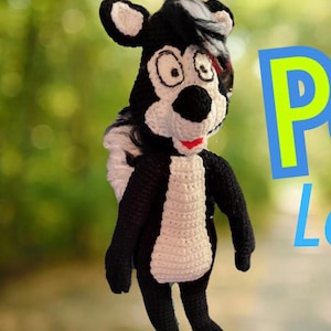 Puede incluir: Un juguete de peluche de mofeta de crochet negro y blanco con una boca roja y ojos grandes. El juguete está de pie sobre un fondo verde y marrón.