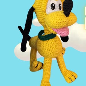 Puede incluir: Un perro de peluche de crochet con pelaje amarillo, orejas y nariz negras, y un collar verde. El perro tiene la lengua rosa que sobresale y está de pie sobre un fondo azul claro.