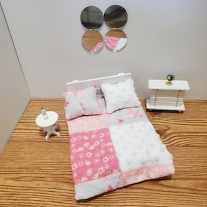 May include: Miniature doll bed with a pink and white patchwork quilt, two pillows, and a white headboard. The quilt features floral and polka dot patterns. A small white table and dresser are also visible.