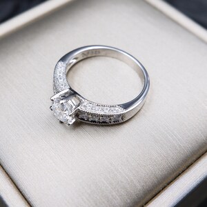 May include: A silver engagement ring with a round, clear center stone and smaller stones set in the band. The ring is displayed in a white box.