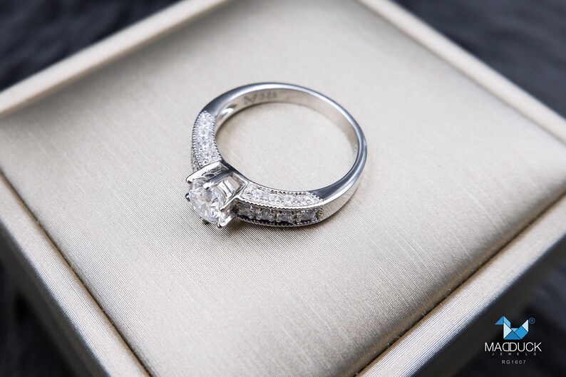 May include: A silver engagement ring with a round, clear center stone and smaller stones set in the band. The ring is displayed in a white box.