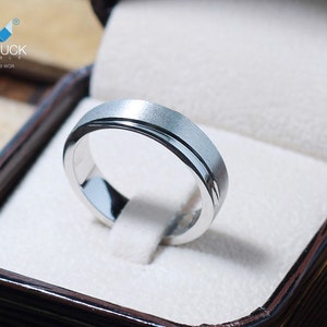 May include: A silver ring with a brushed finish and a polished inner band, presented in a brown jewelry box. The ring features the text "MACDUCK JEWELS" and "RG1728WGH".
