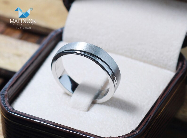 May include: A silver ring with a brushed finish and a polished inner band, presented in a brown jewelry box. The ring features the text "MACDUCK JEWELS" and "RG1728WGH".