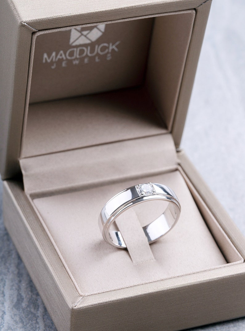 May include: A silver ring with a single diamond set in a white gold band. The ring is in a beige velvet ring box with the words "Madduck Jewels" printed on the inside.