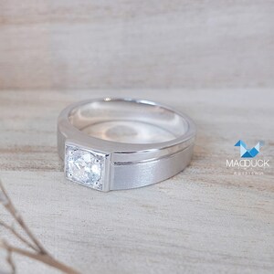 May include: A silver ring with a single, clear, square-shaped gemstone set in the center. The ring has a thick band with a subtle groove.