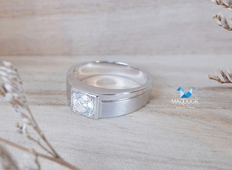 May include: A silver ring with a single, clear, square-shaped gemstone set in the center. The ring has a thick band with a subtle groove.
