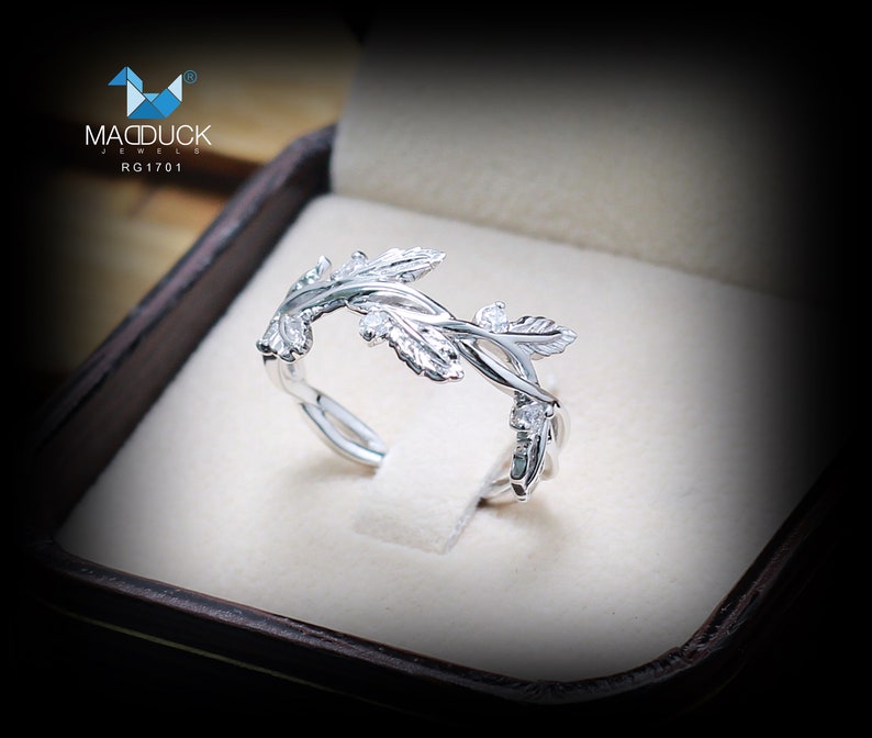 May include: A silver ring with a delicate vine design featuring leaves and small stones. The ring is displayed in a brown box with the text "MACDUCK JEWELS RG1701".