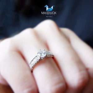 May include: A silver ring with a large, clear, round diamond in the center and smaller diamonds surrounding it. The ring is on a person's finger.