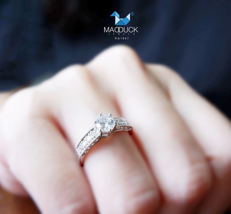 May include: A silver ring with a large, clear, round diamond in the center and smaller diamonds surrounding it. The ring is on a person's finger.