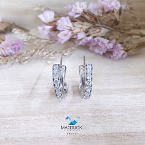 May include: Silver hoop earrings with a row of clear cubic zirconia stones around the edge. The earrings are on a light wood surface.