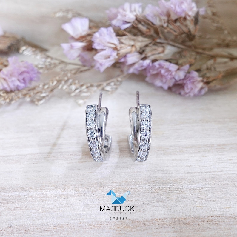 May include: Silver hoop earrings with a row of clear cubic zirconia stones around the edge. The earrings are on a light wood surface.