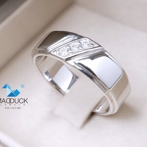 May include: A silver band with a diagonal row of three small diamonds. The band has a polished finish and a slightly rounded edge. The text "MADDUCK JEWELS RG1507WG" is visible in the lower left corner of the image.