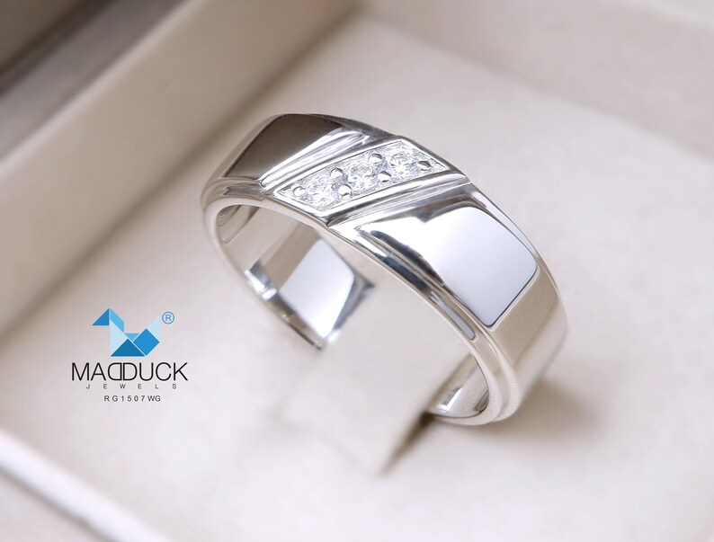 May include: A silver band with a diagonal row of three small diamonds. The band has a polished finish and a slightly rounded edge. The text "MADDUCK JEWELS RG1507WG" is visible in the lower left corner of the image.
