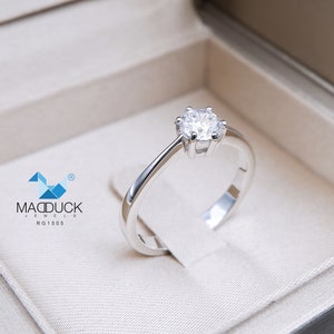 May include: A silver solitaire engagement ring with a round brilliant cut diamond in a white box. The ring is set in a simple band with a four-prong setting. The box has the text "MADDUCK JEWELS RG1505" printed on the inside.
