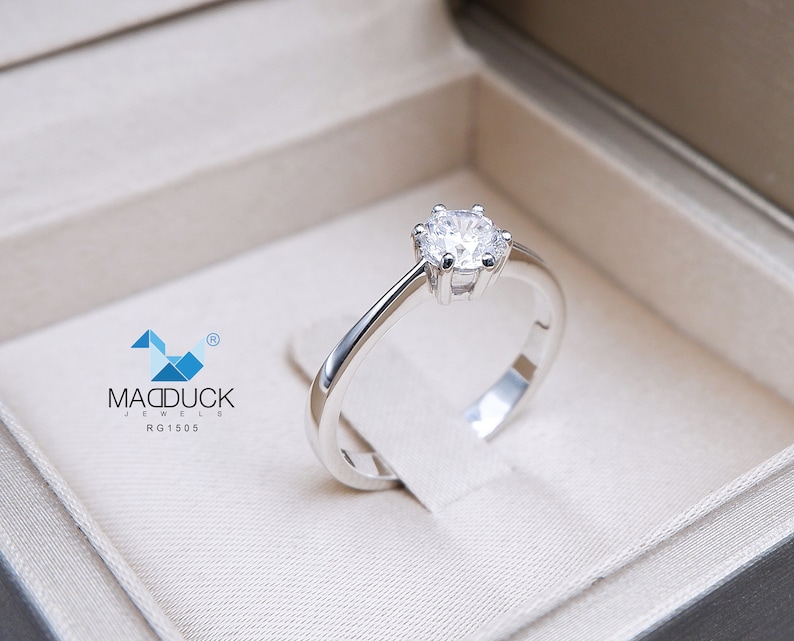 May include: A silver solitaire engagement ring with a round brilliant cut diamond in a white box. The ring is set in a simple band with a four-prong setting. The box has the text "MADDUCK JEWELS RG1505" printed on the inside.