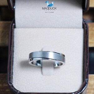 May include: A silver-toned ring with a brushed finish and a diagonal groove, displayed in a brown jewelry box. The ring is set against a white background. The box has the text "MACDUCK JEWELS RG1728WGR".