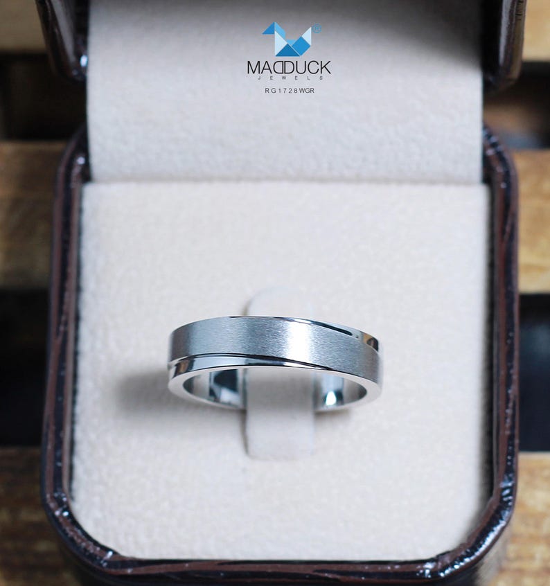 May include: A silver-toned ring with a brushed finish and a diagonal groove, displayed in a brown jewelry box. The ring is set against a white background. The box has the text "MACDUCK JEWELS RG1728WGR".