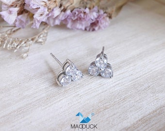 May include: A pair of silver stud earrings with three clear cubic zirconia stones each, arranged in a triangular shape. The earrings are on a white wooden surface.