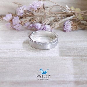 May include: A silver ring with a brushed finish. The ring has a flat top and a slightly raised outer edge. The ring is set against a light wooden surface with dried purple flowers in the background. The ring is from MacDuck Jewels, RG1728 WGR.