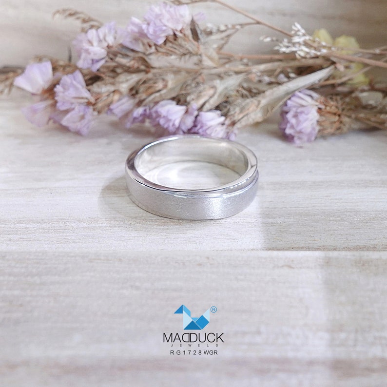 May include: A silver ring with a brushed finish. The ring has a flat top and a slightly raised outer edge. The ring is set against a light wooden surface with dried purple flowers in the background. The ring is from MacDuck Jewels, RG1728 WGR.