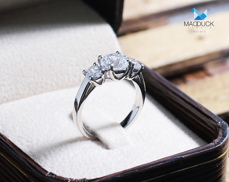 May include: A silver three-stone engagement ring with round diamonds set in a classic prong setting. The ring is displayed in a brown velvet box with the text "MacDuck Jewels RG1415".