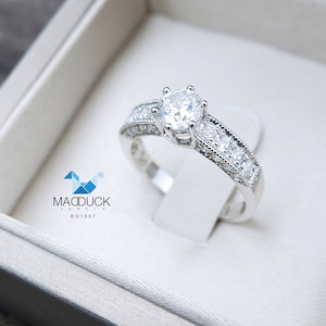 May include: A silver engagement ring with a large, round, clear center stone and smaller clear stones set in a band. The ring is in a white box with the text "MADDUCK JEWELS RG1607".