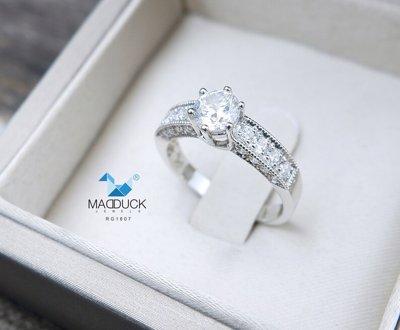 May include: A silver engagement ring with a large, round, clear center stone and smaller clear stones set in a band. The ring is in a white box with the text "MADDUCK JEWELS RG1607".