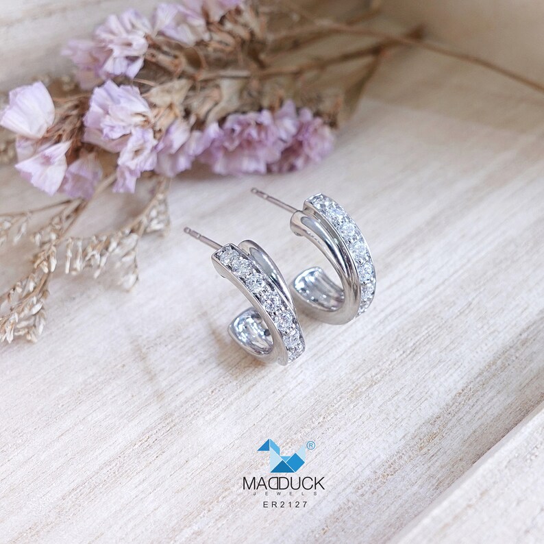 May include: A pair of silver hoop earrings with a double row of clear cubic zirconia stones. The earrings are on a light wood background.
