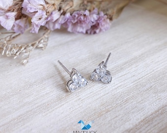 May include: A pair of silver stud earrings with three clear cubic zirconia stones each, arranged in a triangular shape. The earrings are on a white wooden surface.