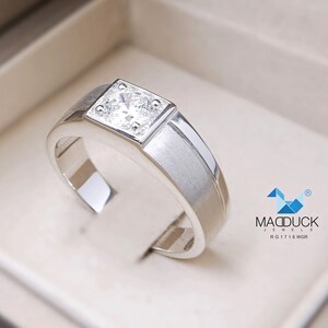 May include: A silver ring with a square-shaped setting featuring a clear, round gemstone. The ring has a brushed finish and a beveled edge. The ring is displayed in a white box with the text "MADDUCK JEWELS RG 17 16 WGR" printed on the bottom.