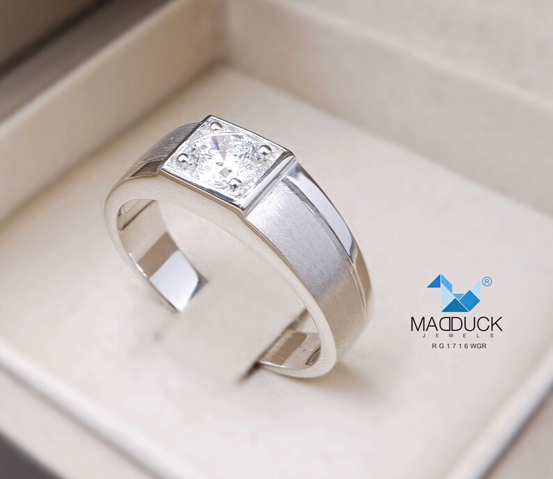 May include: A silver ring with a square-shaped setting featuring a clear, round gemstone. The ring has a brushed finish and a beveled edge. The ring is displayed in a white box with the text "MADDUCK JEWELS RG 17 16 WGR" printed on the bottom.