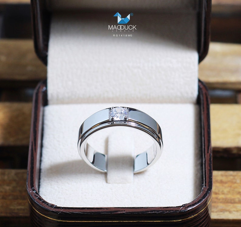 May include: A silver wedding band with a single, round diamond set in the center. The band has a grooved design and is displayed in a brown box.