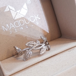 May include: A silver ring with a delicate design of intertwined branches and leaves, accented with small clear stones. The ring is displayed in a beige jewelry box with the brand name "MADDUCK JEWELS" printed on the lid.