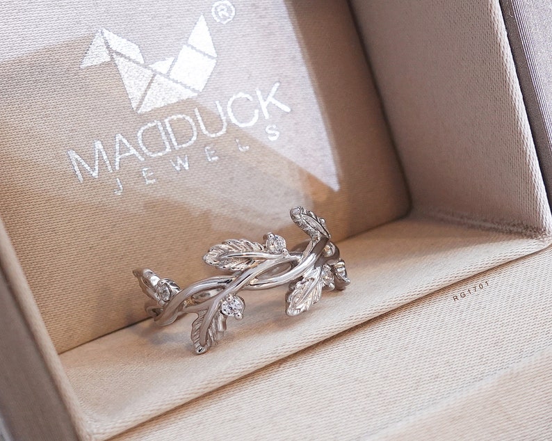 May include: A silver ring with a delicate design of intertwined branches and leaves, accented with small clear stones. The ring is displayed in a beige jewelry box with the brand name "MADDUCK JEWELS" printed on the lid.