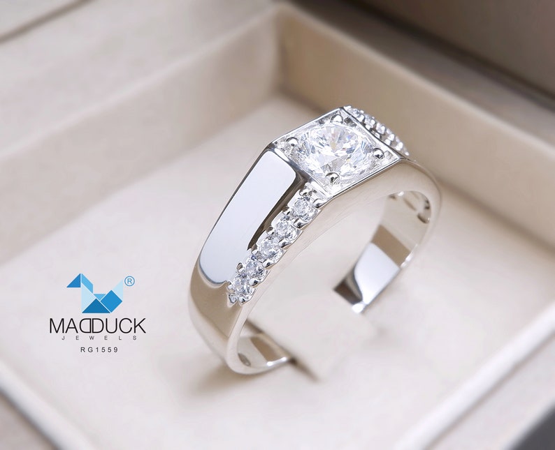 May include: A silver ring with a large, clear, round gemstone surrounded by smaller, clear gemstones. The ring is set in a square-shaped band.